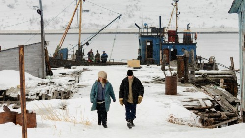 A warmly-dressed man and woman walk in snow towards the viewer. Behind them is a snowy jetty where a large battered fishing boat is moored. A snowy mountain can be seen in the distance.