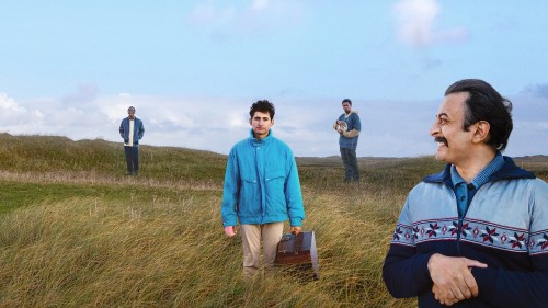 In the centre of the image stands a man in a blue coat carrying an instrument case. Two other men stand in the background. Another man stands on the right, arms folded, grinning, and looking back towards the man in blue. Overall scenery is windswept, grassy hillside.