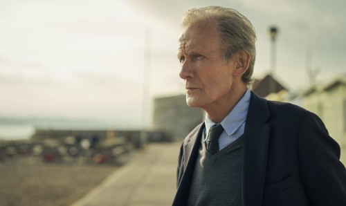 A smartly dressed older man looks towards the left. A beach can be seen out of focus in the background.