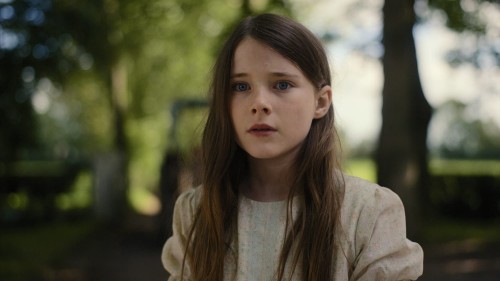 A young girl with long brown hair faces slightly away from the viewer. A woodland clearing can be seen in the background.